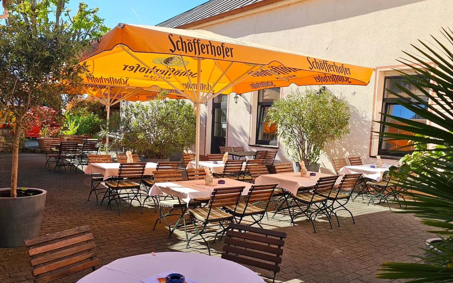 Weiß eingedeckte Tische unter orangefarbenen Sonnenschirmen im Biergarten